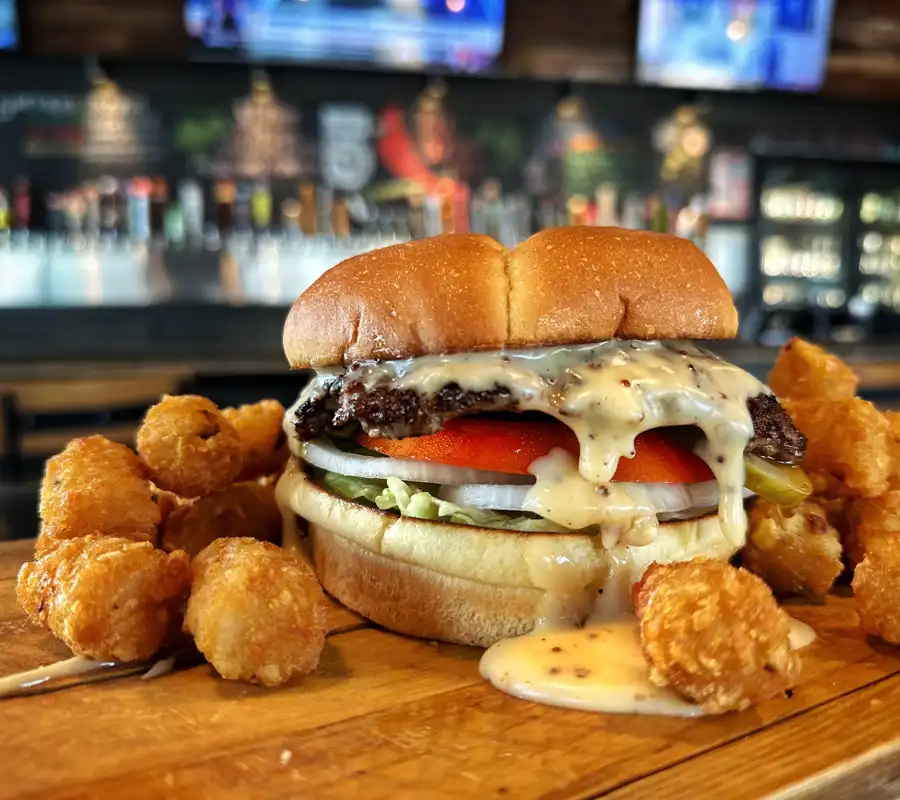 smash burger with dripping beer cheese tomato onion lettuce on a bun surrounded by tater tots